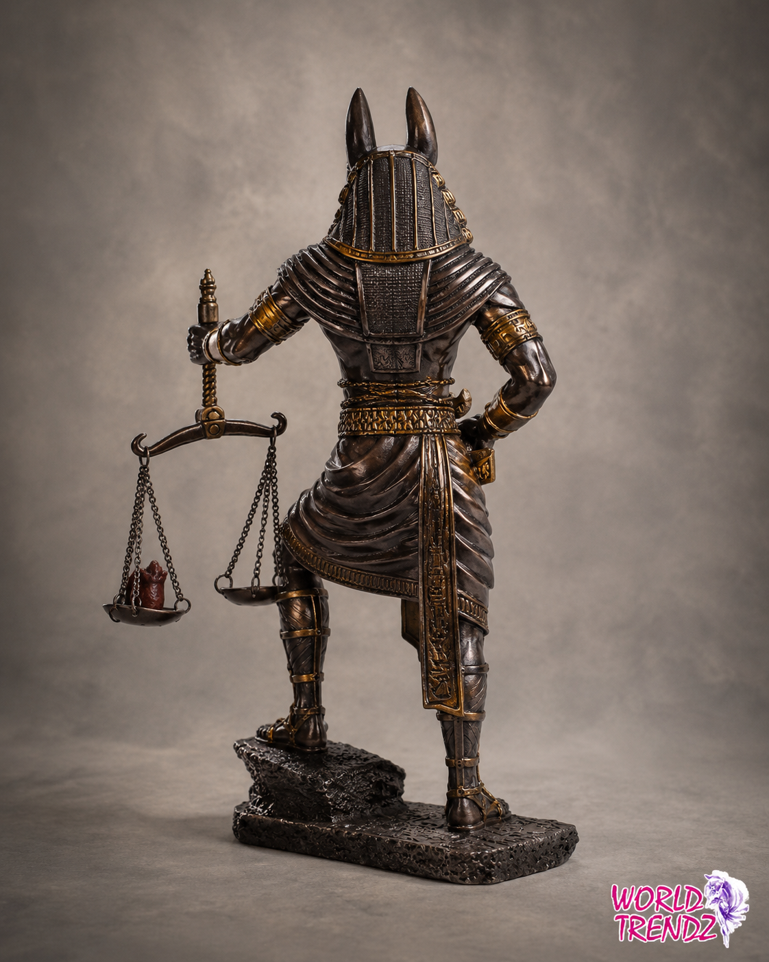 A detailed statue of Anubis, the Egyptian god of justice, shown from the back. He is depicted in dark metallic armor with gold accents, holding a scale of justice in one hand, standing on a dark textured base against a gray studio background.