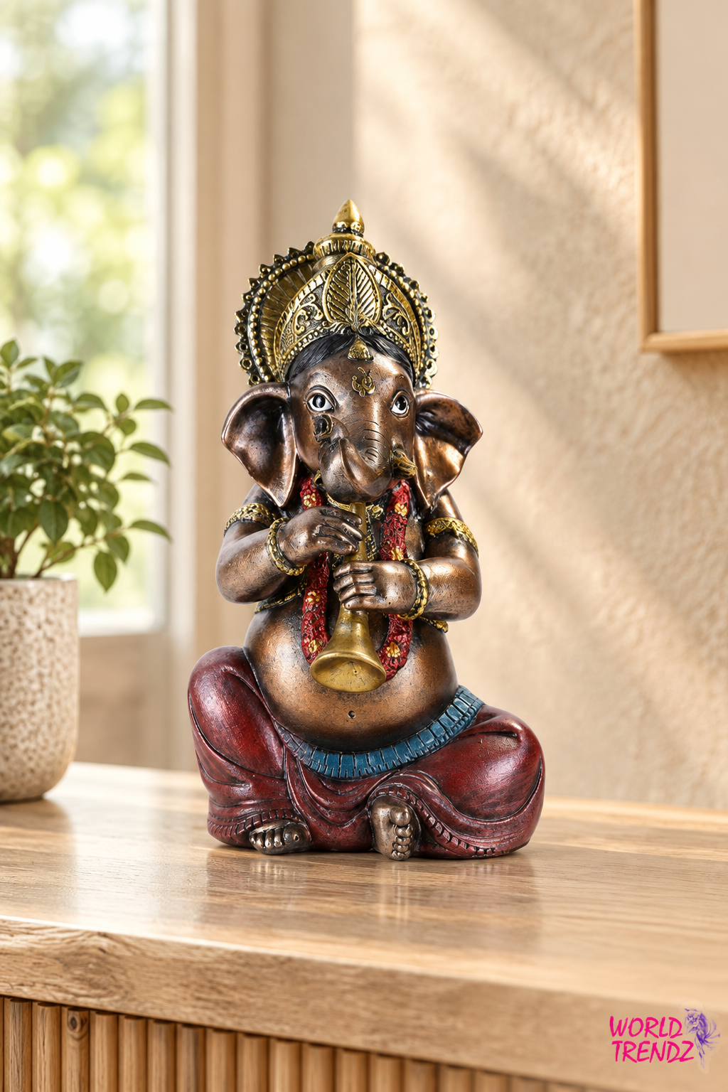 A decorative statue of Ganesha seated on a wooden surface in a warm indoor setting. The statue has a metallic bronze finish with gold detailing on the crown and belly, red drapery, and a blue waist sash.