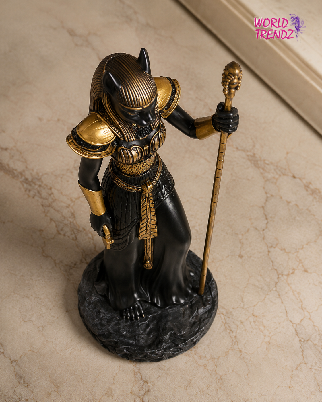 An Egyptian Bastet Goddess Statue, featuring a black body with gold armor details, standing on a dark textured base and holding a gold staff, photographed on a beige marble-patterned floor.