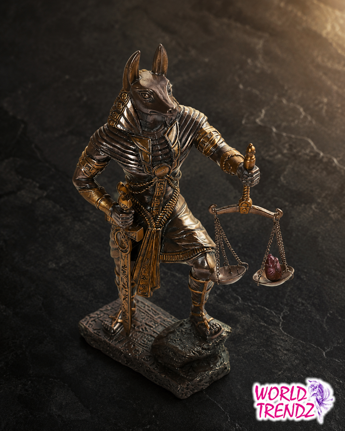 An Egyptian Anubis statue holding a balance scale and a sword, crafted in a metallic gold and bronze finish, standing on a dark textured base against a dark stone background.