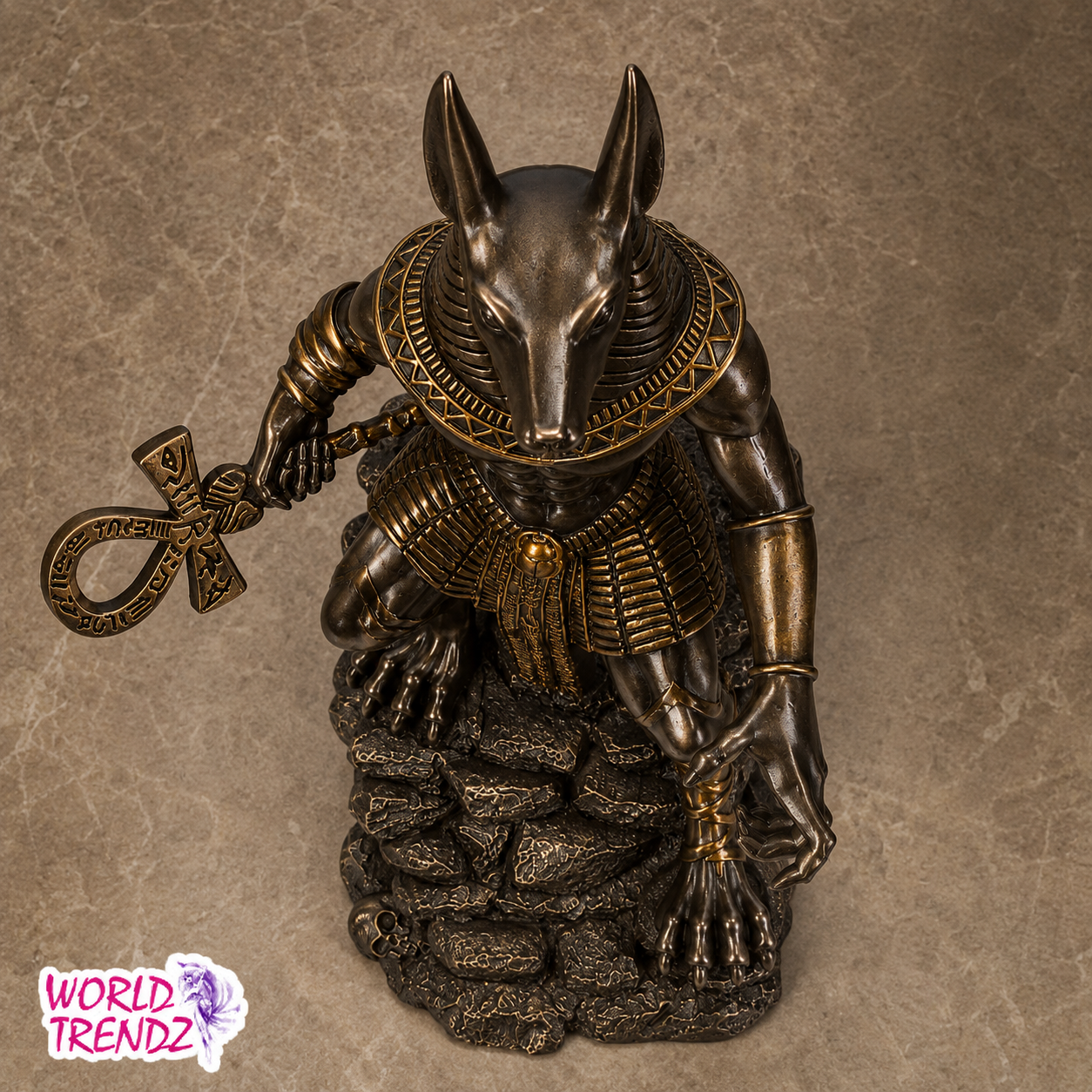 An Egyptian Anubis statue made of dark bronze metal with gold accents, holding an ankh symbol, set against a textured brown background. A 'WORLD TRENDZ' logo is visible in the bottom-left corner.