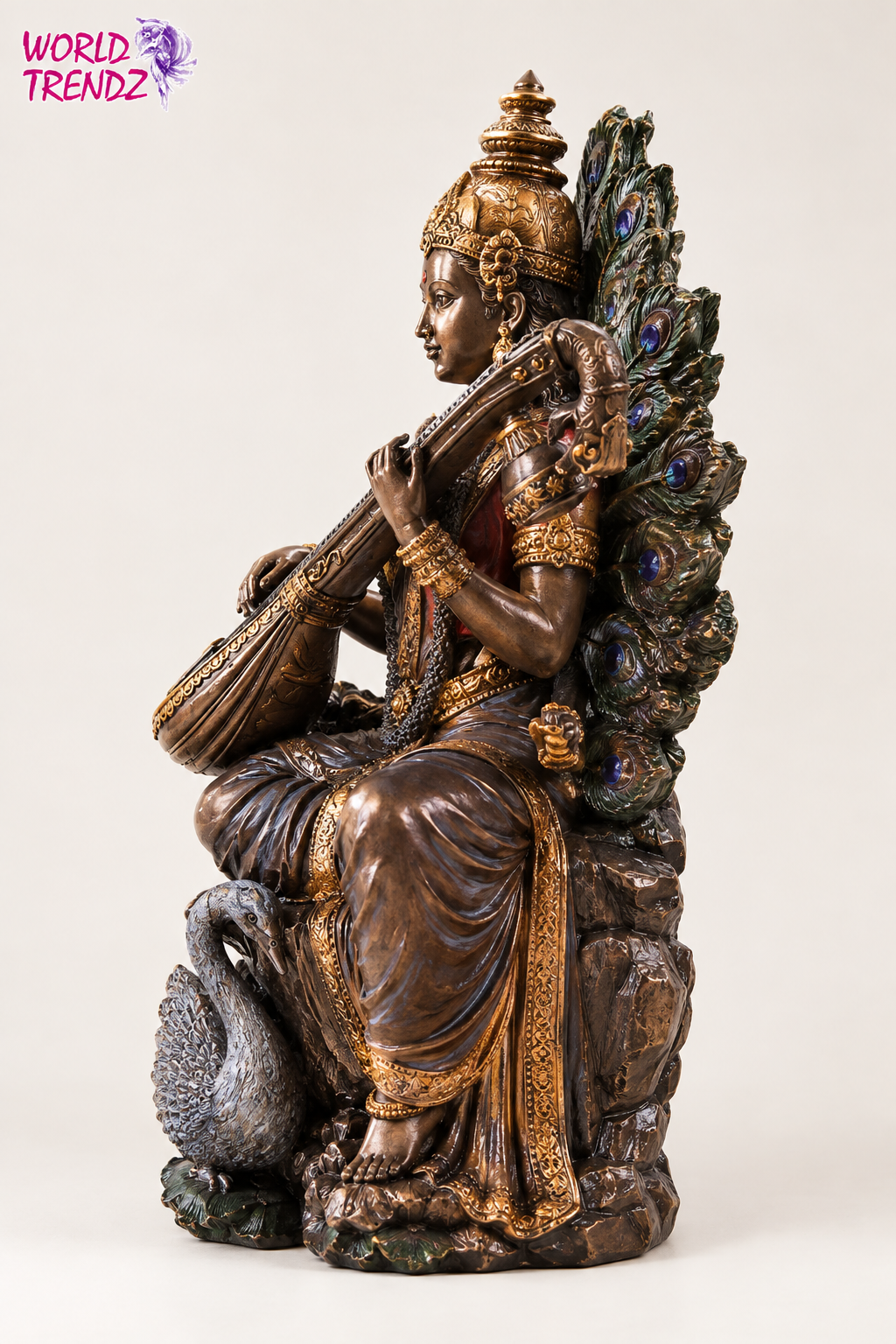 A bronze-finished statue of a seated Goddess Saraswati playing a stringed instrument, adorned with gold detailing and a large peacock feather backrest, shown on a plain light background.