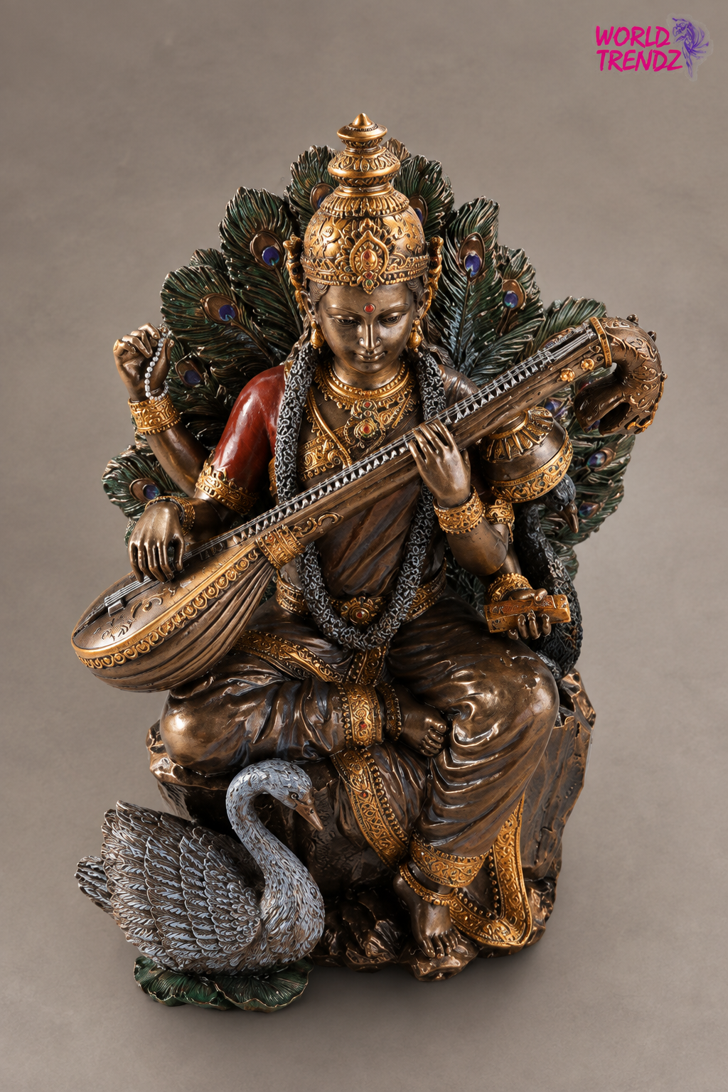 An ornate bronze-finished statue of a seated Hindu deity Goddess Saraswati playing a stringed instrument, decorated with gold detailing, a peacock feather headdress and backrest, and a swan at the base, photographed against a plain taupe background.