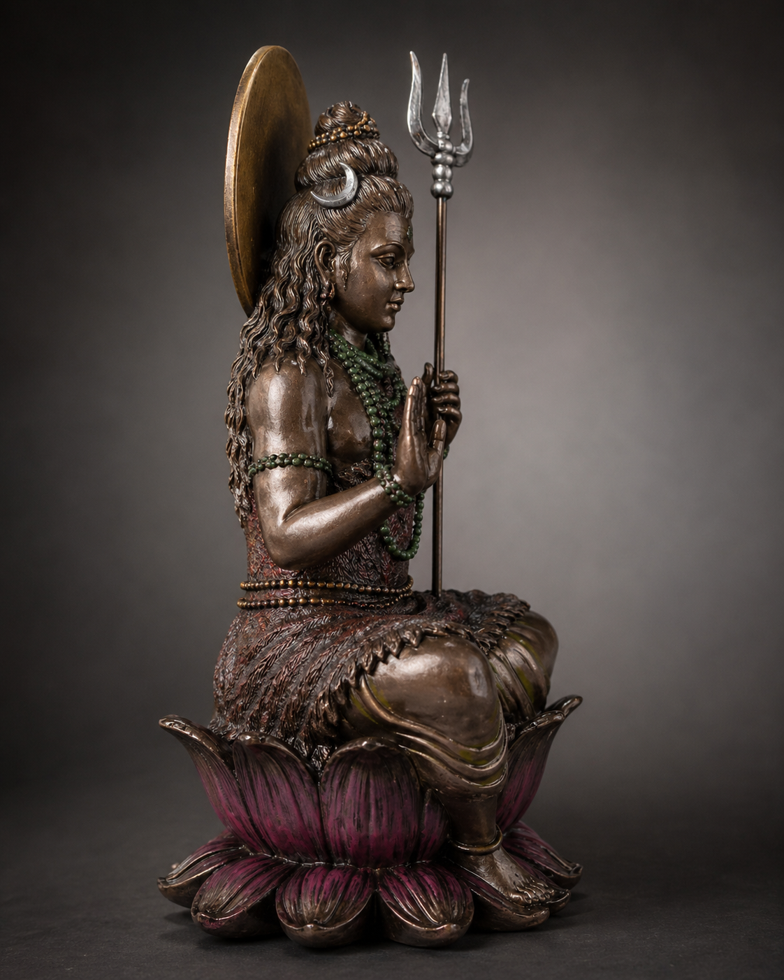 Bronze statue of seated Shiva holding a trident on a lotus flower against a gray background
