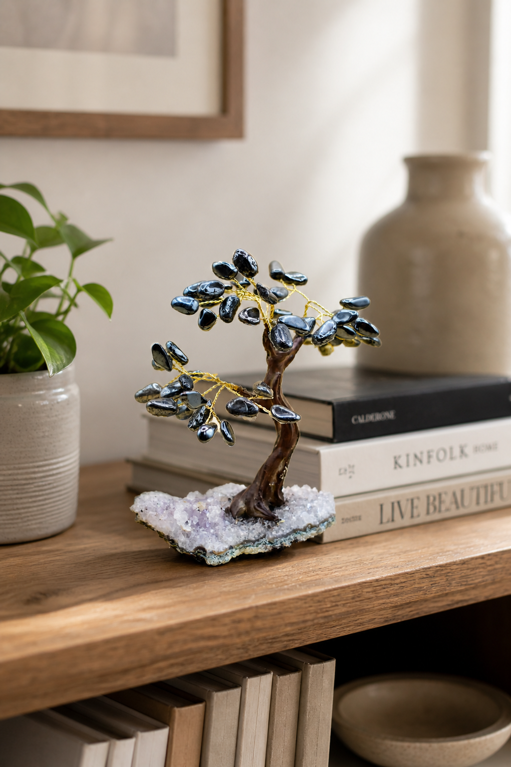 The hematite gemstone tree placed on a styled living room shelf with stacked books, a small potted plant, and minimalist decor items. 