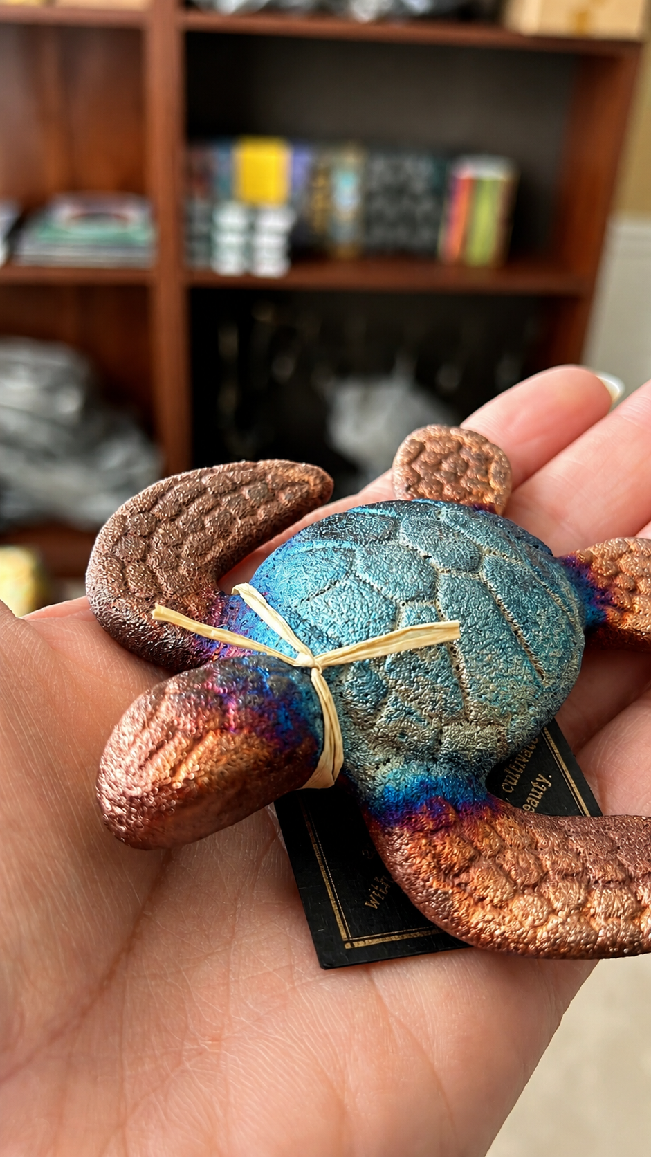 Hand holding a small, colorful turtle figurine with a blurred bookshelf in the background