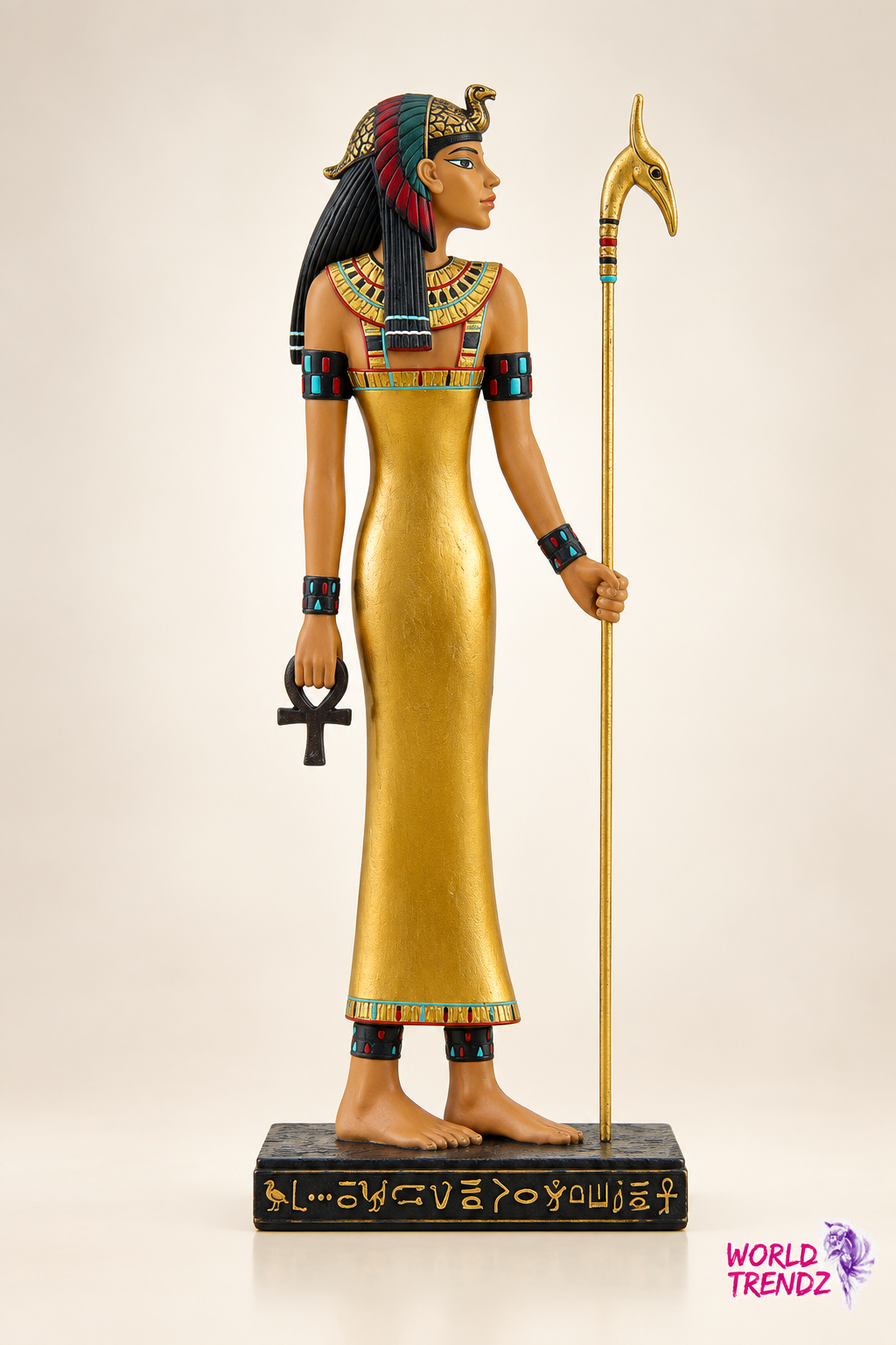 An Egyptian goddess figurine Isis standing on a black base, wearing a long gold dress and headdress, holding an ankh and a staff, photographed against a plain light beige background.