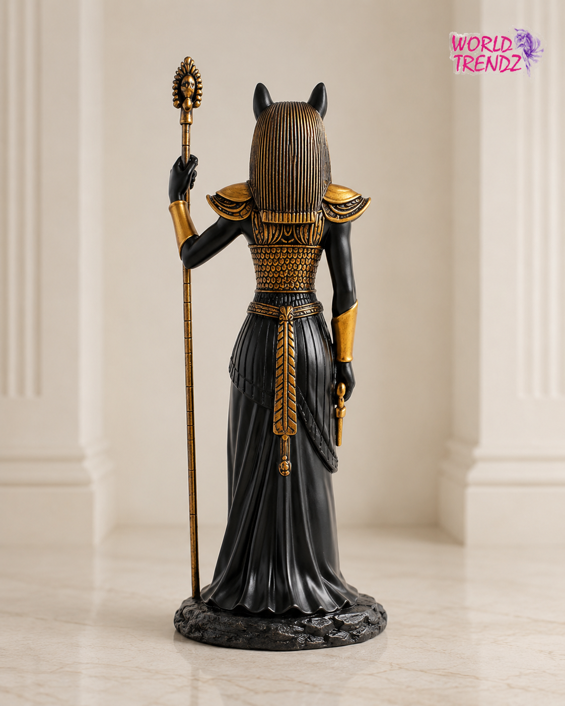 An Egyptian Bastet Goddess Statue shown from the back, featuring a black body, gold headdress and armor details, holding a gold staff, set against a neutral cream-colored indoor background.
