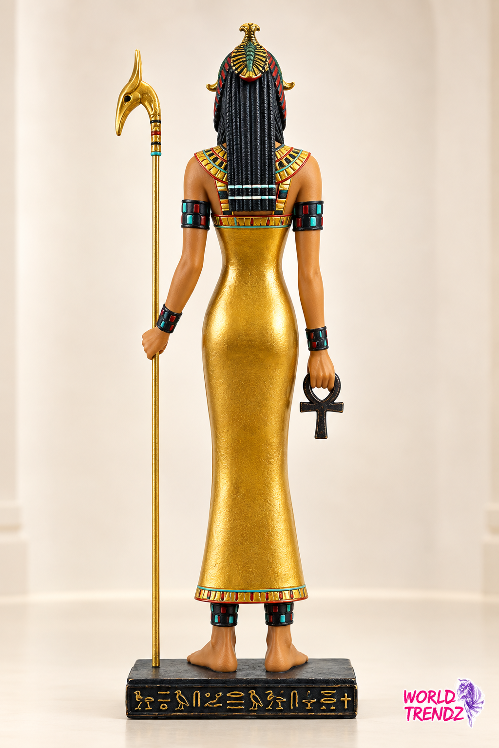 An Egyptian goddess figurine Isis shown from the back, wearing a gold dress and headdress, holding a staff and ankh, standing on a black base with hieroglyphs.