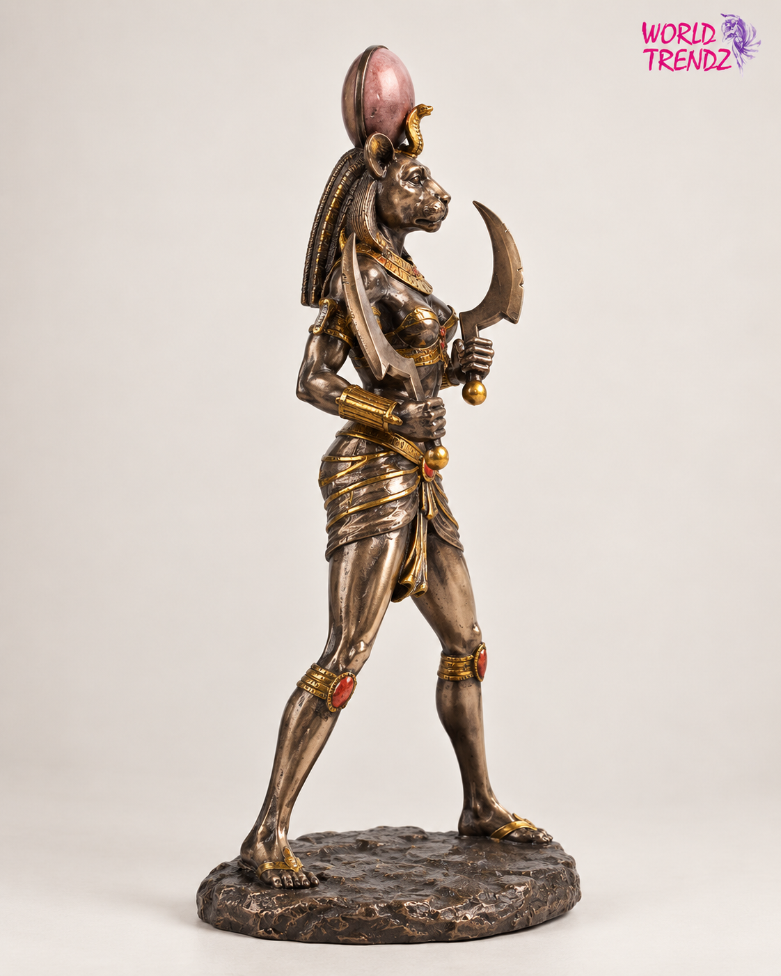 An Egyptian Sekhmet Statue holding a scythe, finished in a metallic bronze finish with gold accents and a pink sun disc on her head, standing on a textured base against a light gray background.