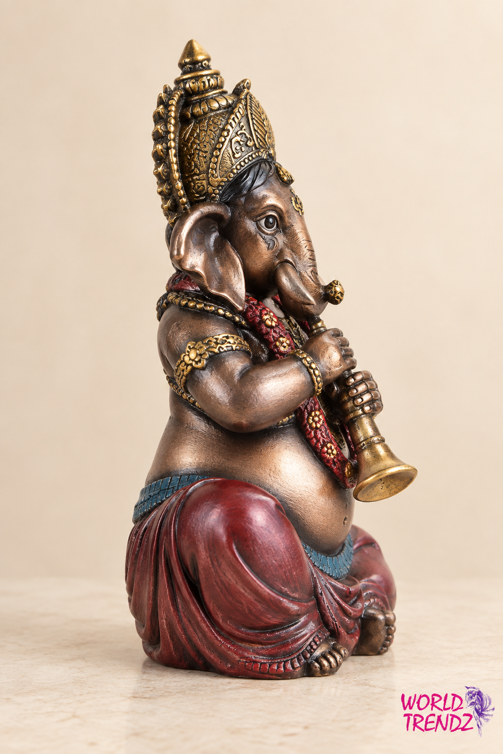 A decorative statue of Lord Ganesha in a seated position, holding a small brass-like instrument. The sculpture has a metallic bronze finish with red draped fabric at the waist and a gold crown and jewelry.