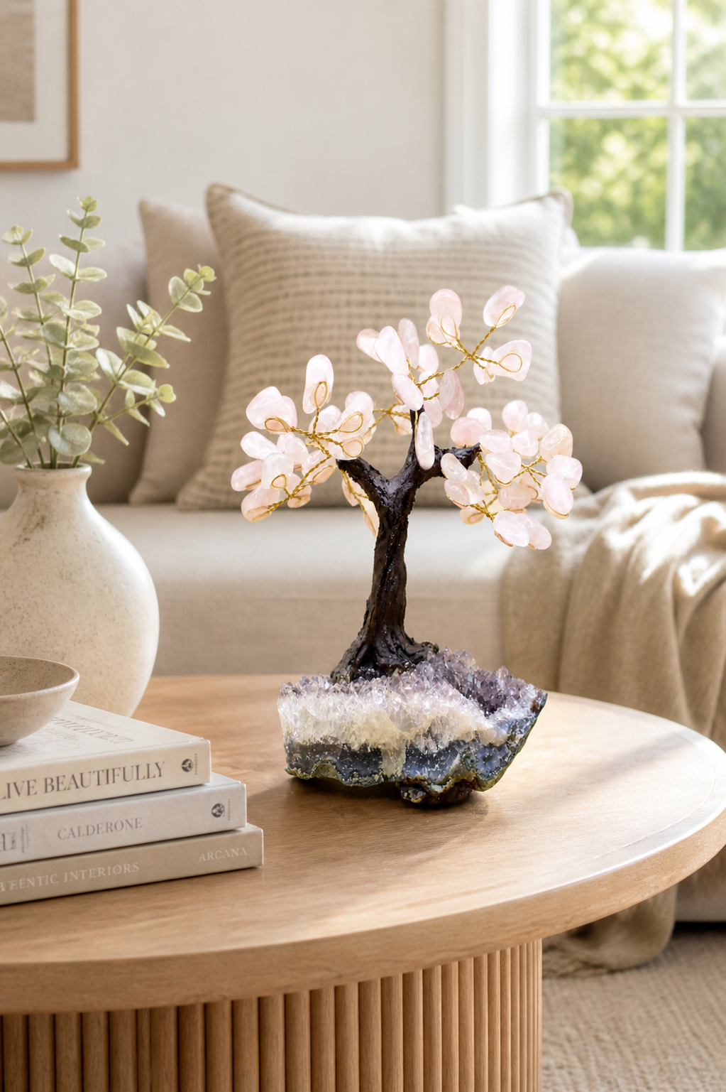 Rose quartz gemstone tree on a modern living room coffee table with soft natural daylight streaming through a nearby window and surrounded with neutral-toned decor such as books, a ceramic vase, and a linen sofa in the background.