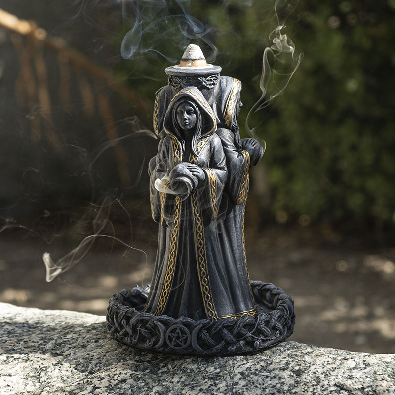 A Mother Maiden Crone Mugga Incense Burner with gold detailing stands on a circular base outdoors, with smoke rising around it and a blurred natural background.