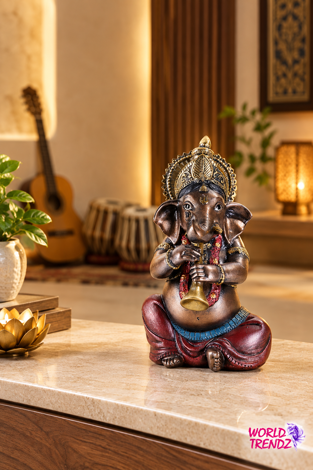 A bronze-finished Lord Ganesha statue seated and holding a flute, placed on a beige marble countertop in a warm, decorated room with a guitar, drum set, and lit lamps in the background.