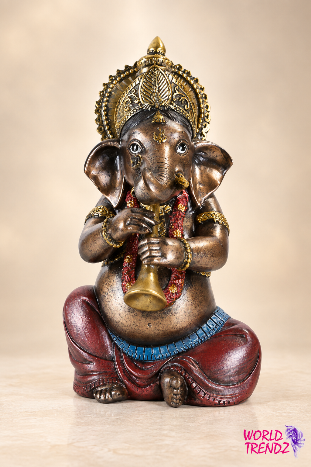 A decorative Ganesha statue with a metallic bronze finish, wearing a gold crown and red draped clothing, seated and holding a brass flute. The statue is photographed against a soft beige studio background with a 'World Trendz' logo in the bottom right corner.