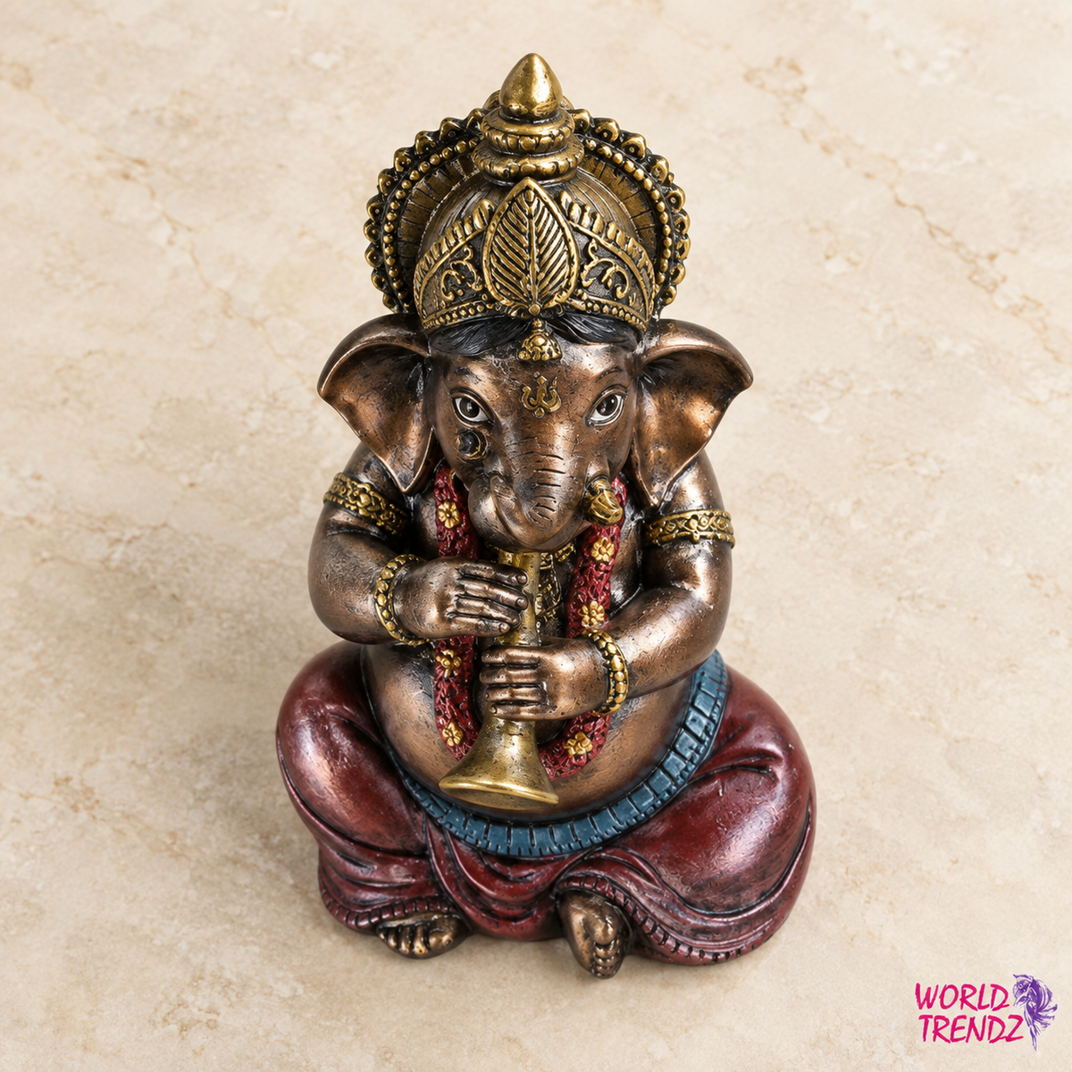 A decorative brass-colored Ganesha statue seated cross-legged, holding a mace and a flute, with red draped clothing and gold ornamental details, photographed on a light beige stone-like surface.