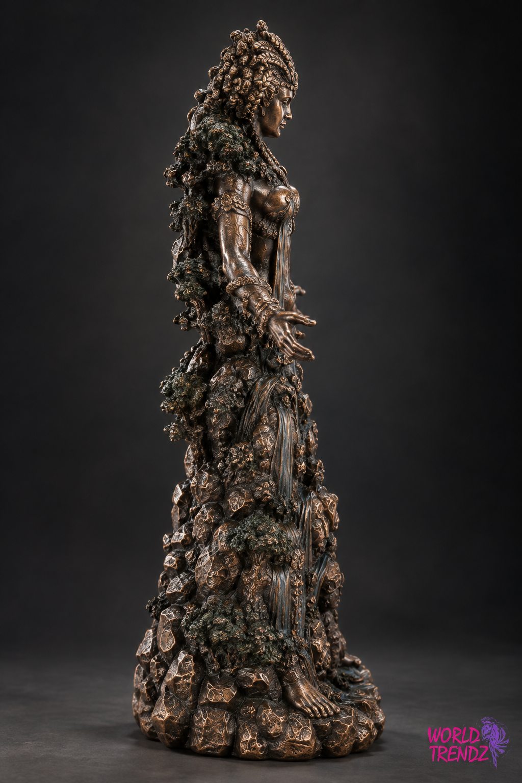 A detailed Goddess Danu with long curly hair, standing amidst rocky, mossy textures. The sculpture has a metallic bronze finish with dark green patina accents, photographed against a dark gray studio background.