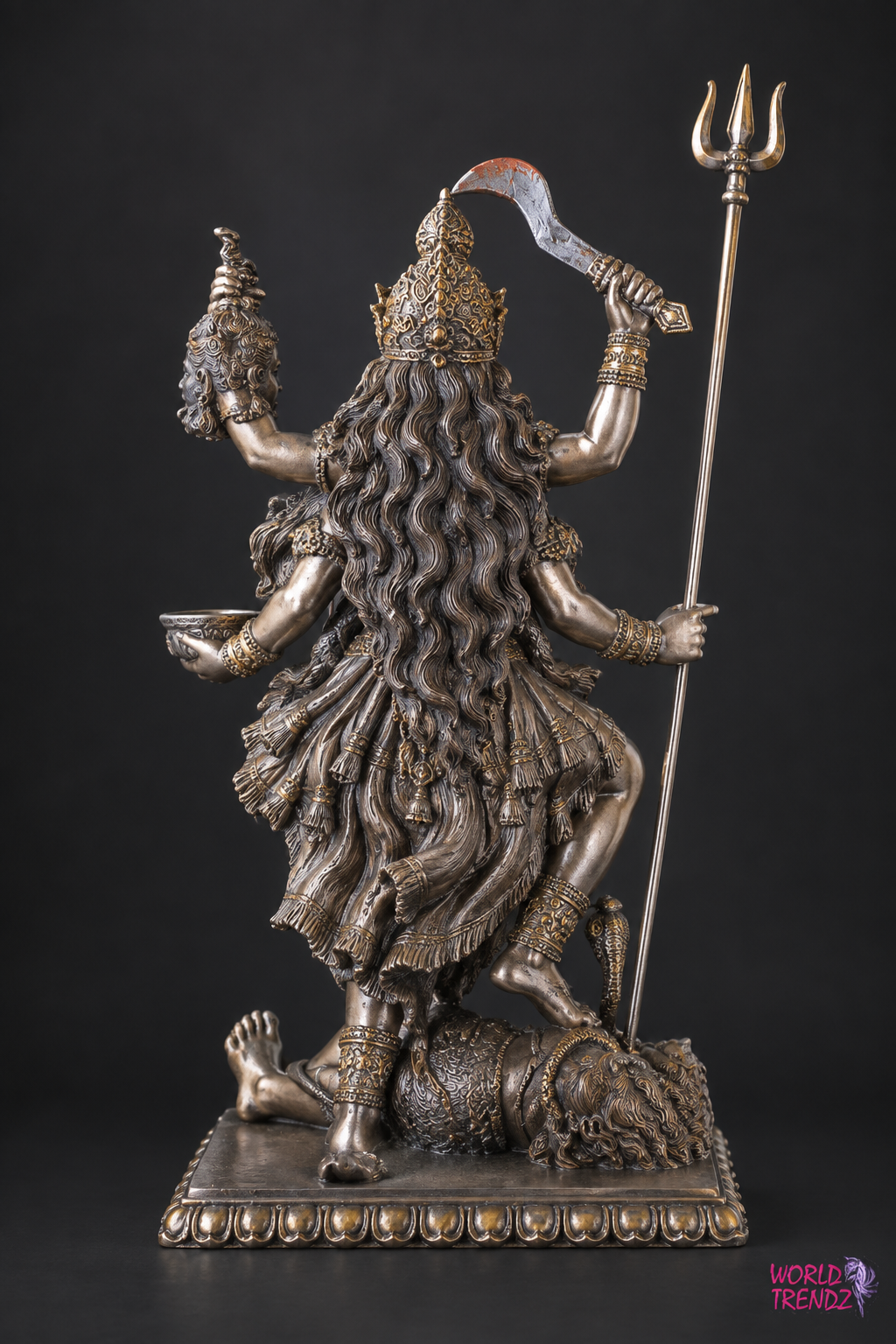 A detailed bronze-finished statue of a Hindu goddess Kala Kali with multiple arms, holding a trident and a curved sword, standing on a base with a defeated figure at her feet, photographed against a solid black background.