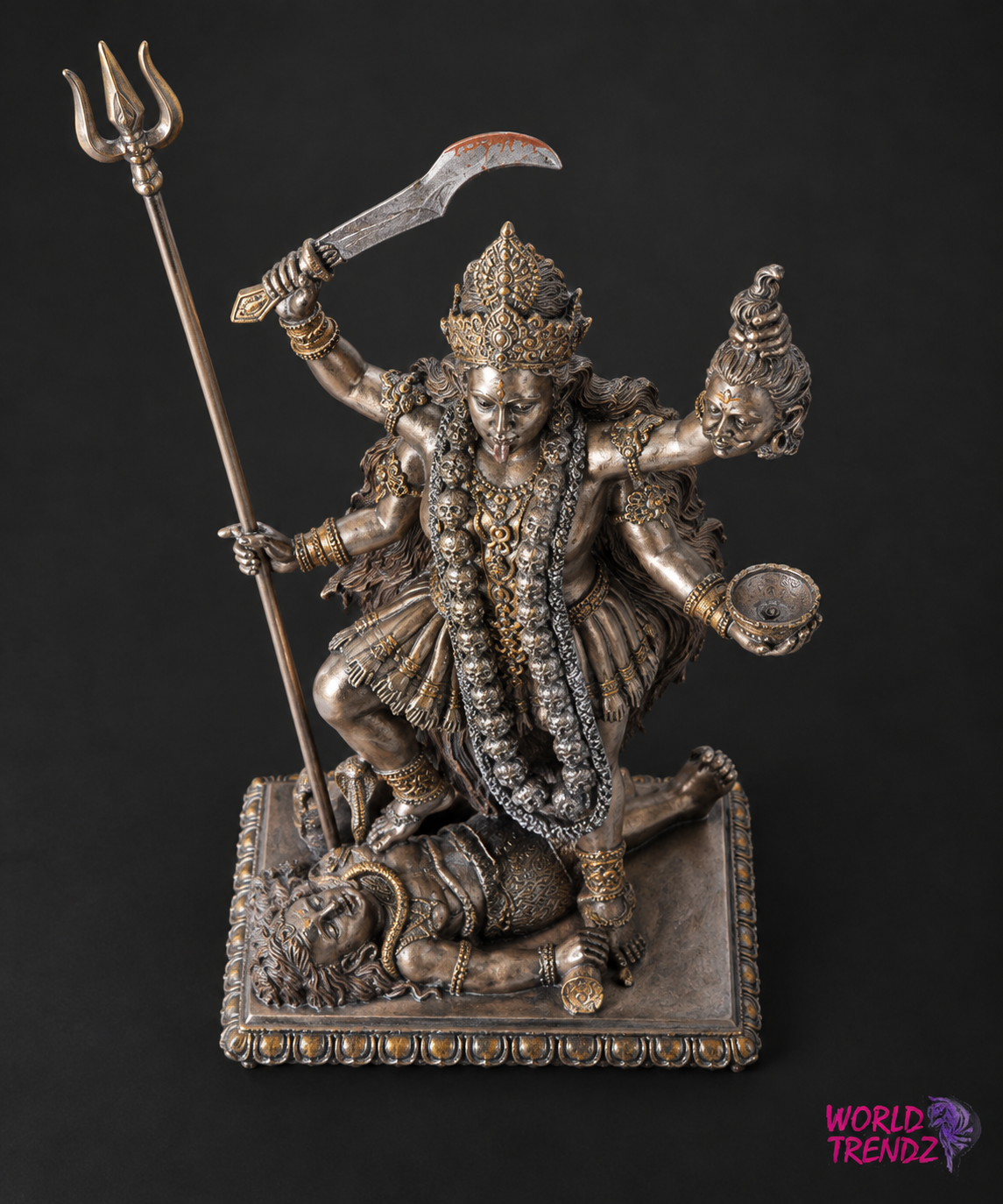 A detailed metallic statue of a Hindu goddess Kala Kali standing on a defeated demon, holding a trident and a curved sword, with ornate jewelry and a decorative base, photographed against a dark background.
