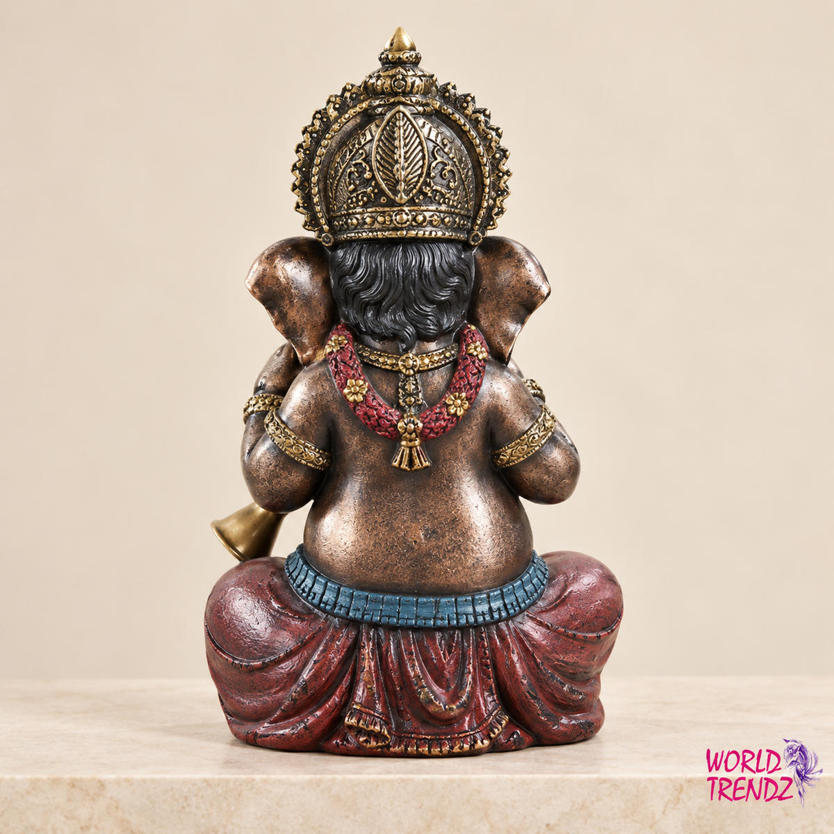 A decorative statue of Ganesha shown from the back, featuring a metallic bronze finish with red draped fabric, a blue waist band, and gold crown and jewelry details, set against a plain beige background.