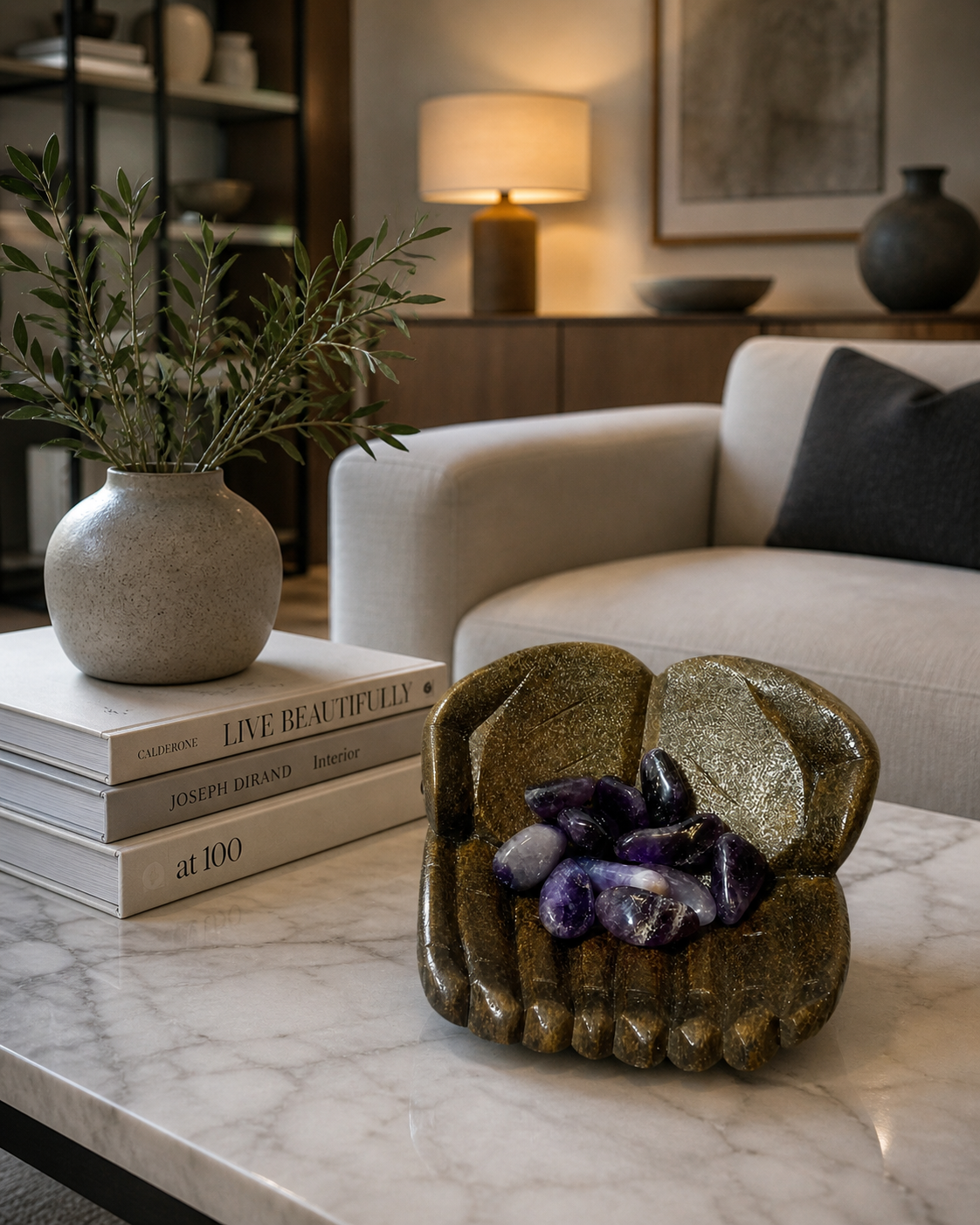 A modern living room interior with neutral tones and elegant styling. The carved bowl with ametrine stones is positioned on a marble coffee table alongside a small stack of design books and a ceramic vase. Soft ambient lighting enhances the texture of the bowl and the gemstones, creating a luxurious and tasteful décor vibe.