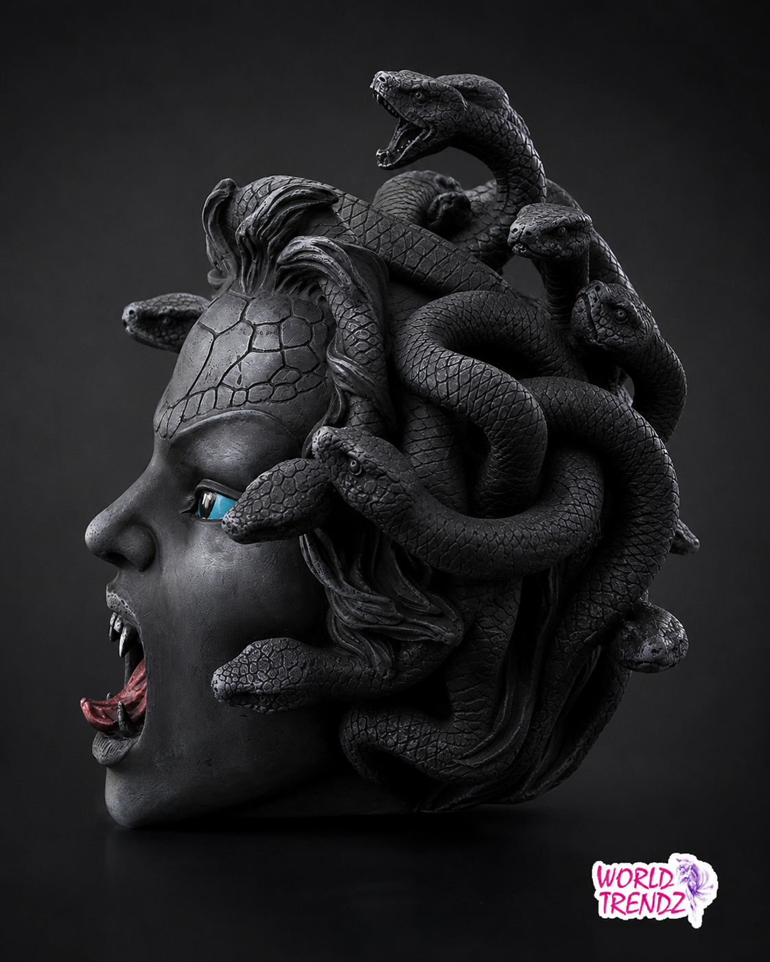 A dark, gothic-style sculpture of The Medusa Head with black snakes coiled through her hair, set against a dark studio background. The sculpture has a cracked stone-like texture on its forehead, a single bright blue eye, and an open mouth showing teeth and red tongue.